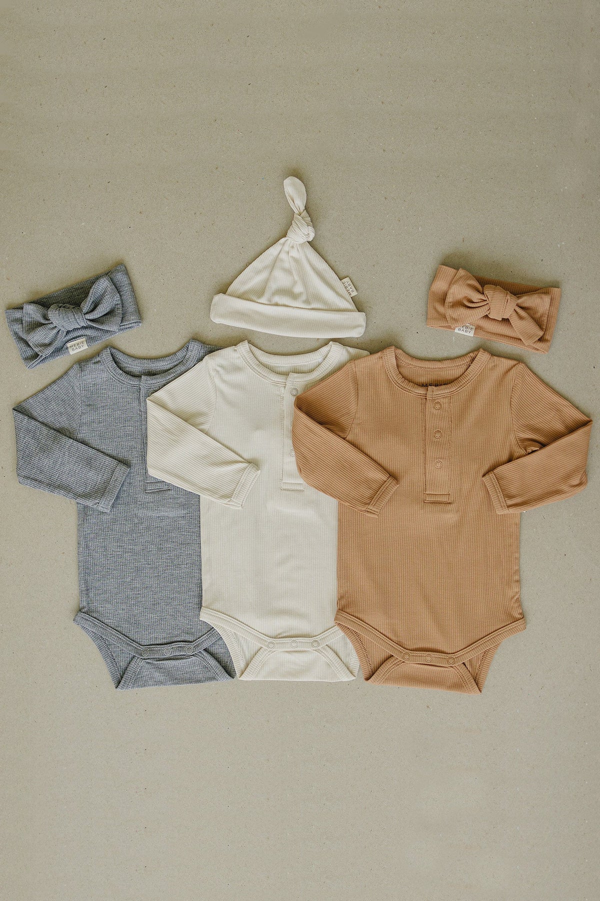 Three ribbed bodysuits in grey, cream, and tan, with matching headbands and a knotted hat displayed on beige.