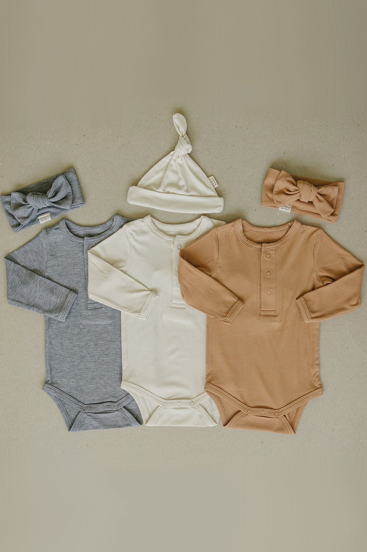 Three ribbed long sleeve bodysuits in gray, white, and brown, with matching headbands and a knotted hat.
