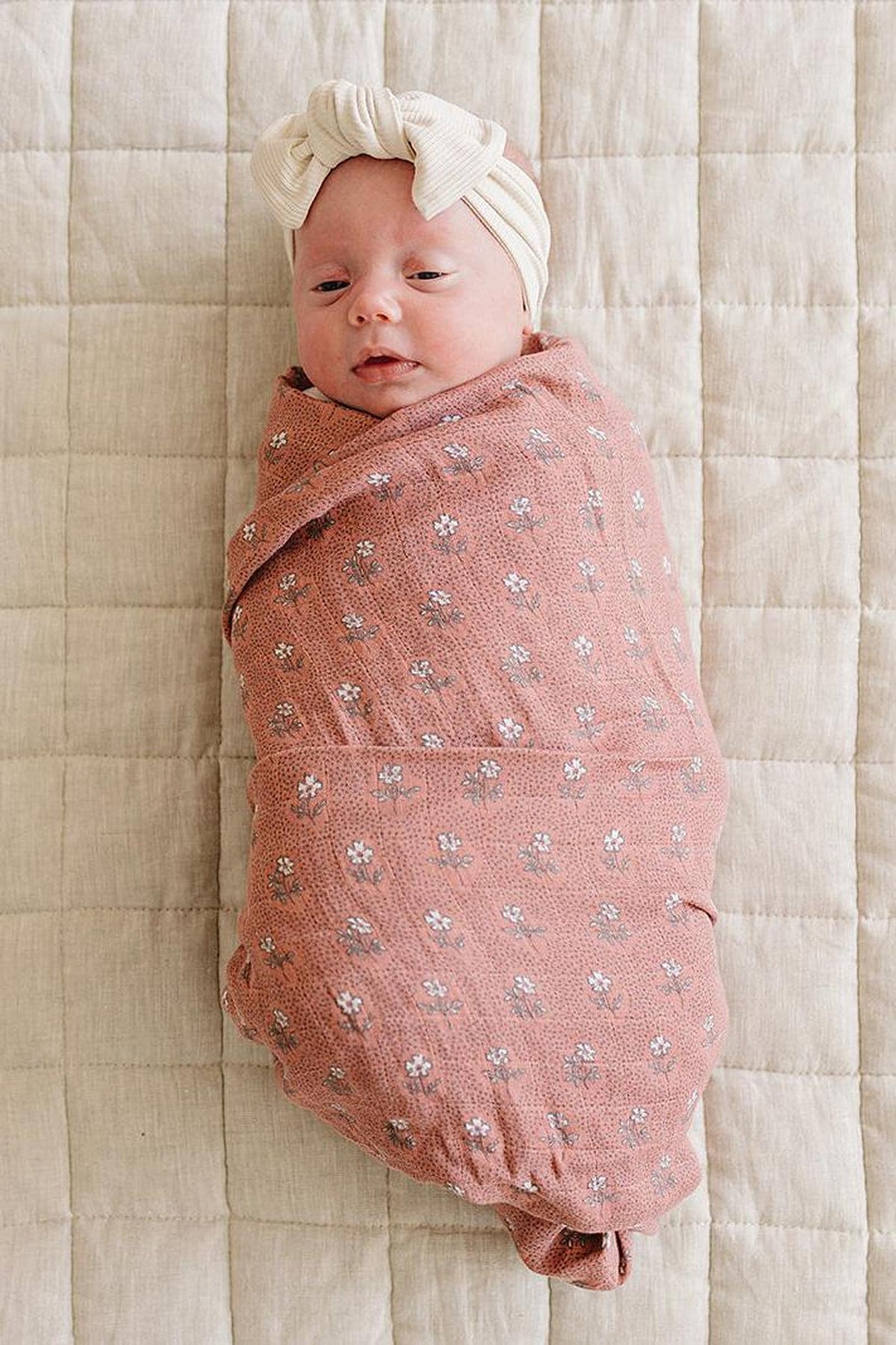 A baby is wrapped in a pink floral muslin swaddle blanket with a cream headband on a quilted surface.