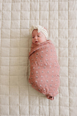 A baby wrapped in a pink floral muslin swaddle blanket lies on a quilted beige surface.