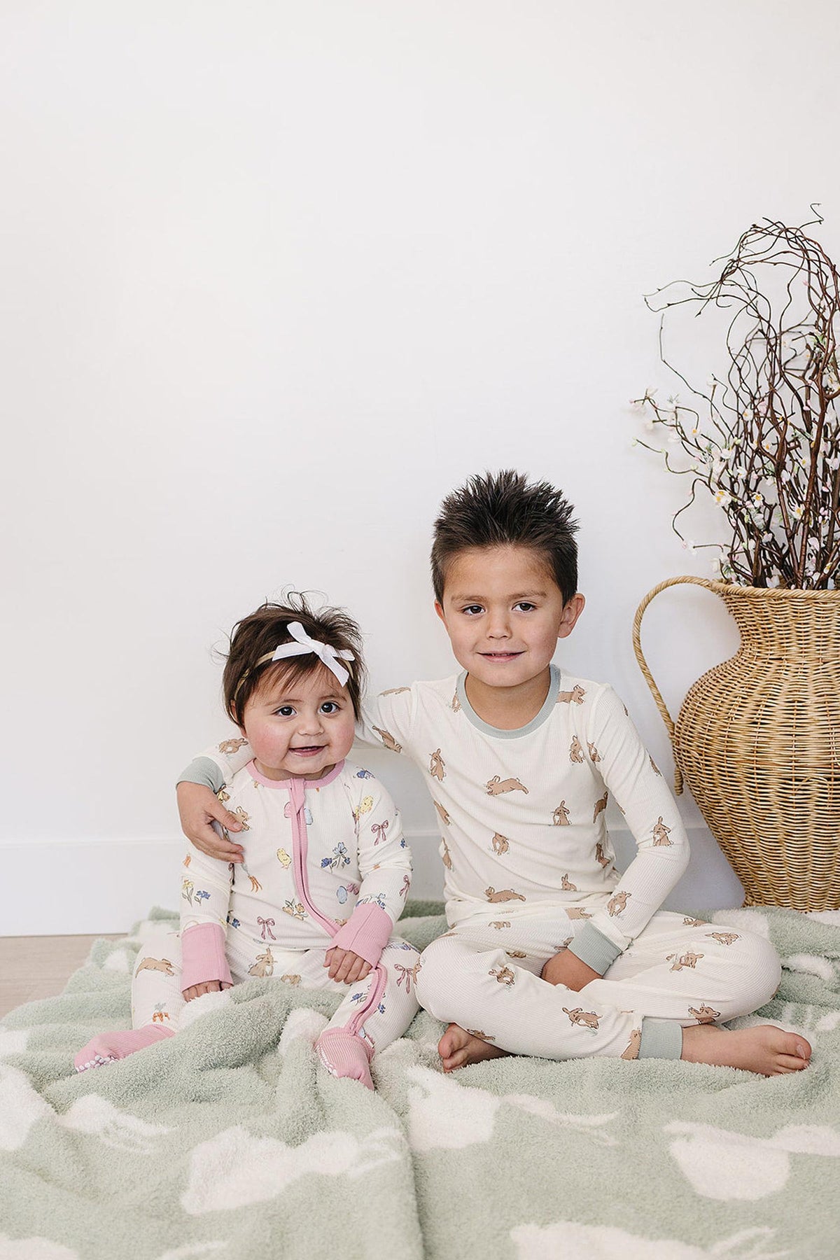 A toddler and a baby sit on a soft, light green blanket, wearing matching ribbed bamboo pajamas featuring adorable bunny prints. The cozy setting highlights their playful, comfortable look.