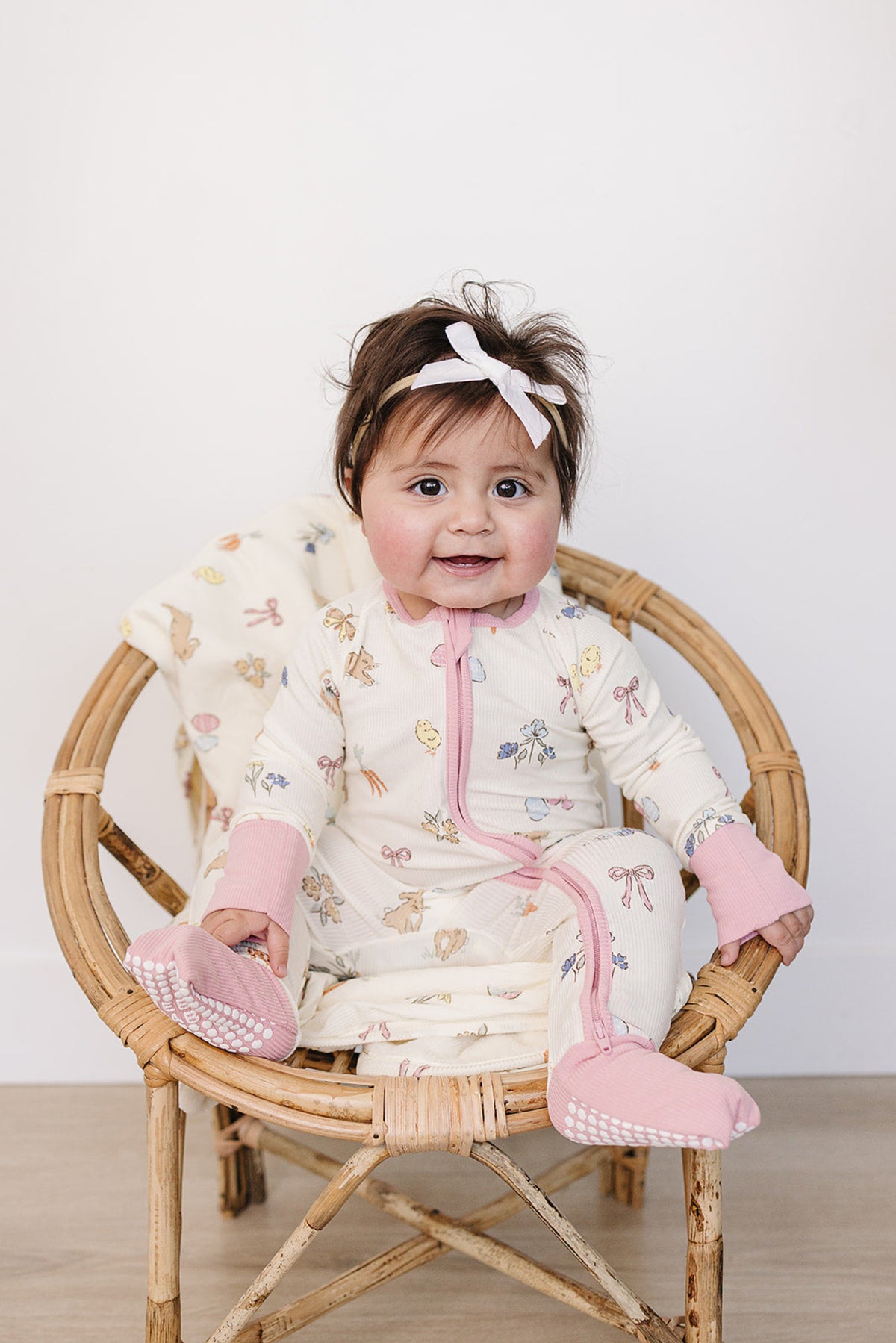 A baby girl sits in a rattan chair, wearing a cream-colored bamboo zipper pajama adorned with pastel floral prints and pink accents, showcasing comfort and style for warmer days.