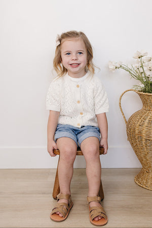 A smiling young girl sits on a wooden stool, wearing a white knitted cardigan, denim shorts, and brown sandals.