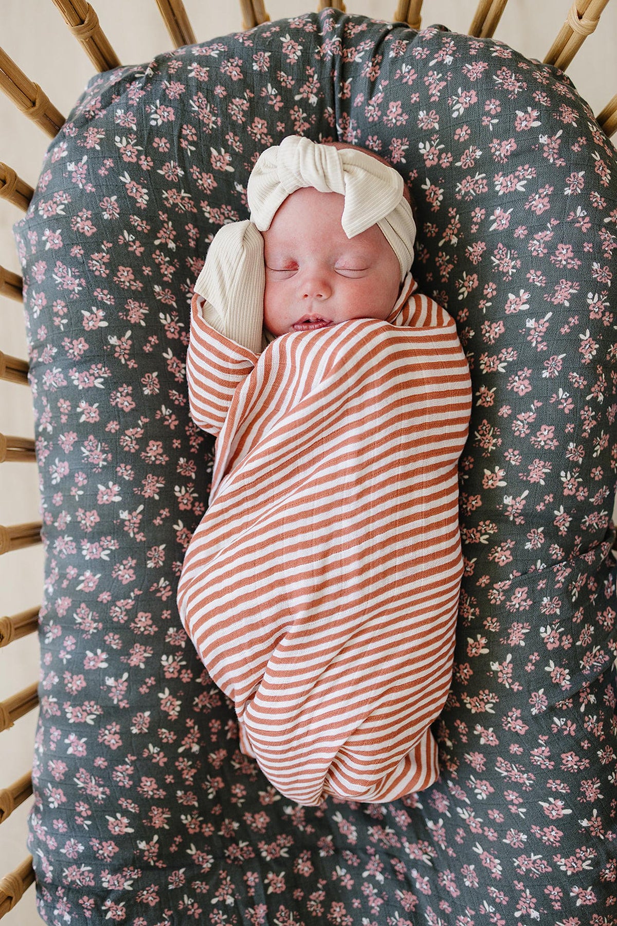 A baby sleeps wrapped in a pink striped muslin swaddle, lying on a floral-patterned cushion.