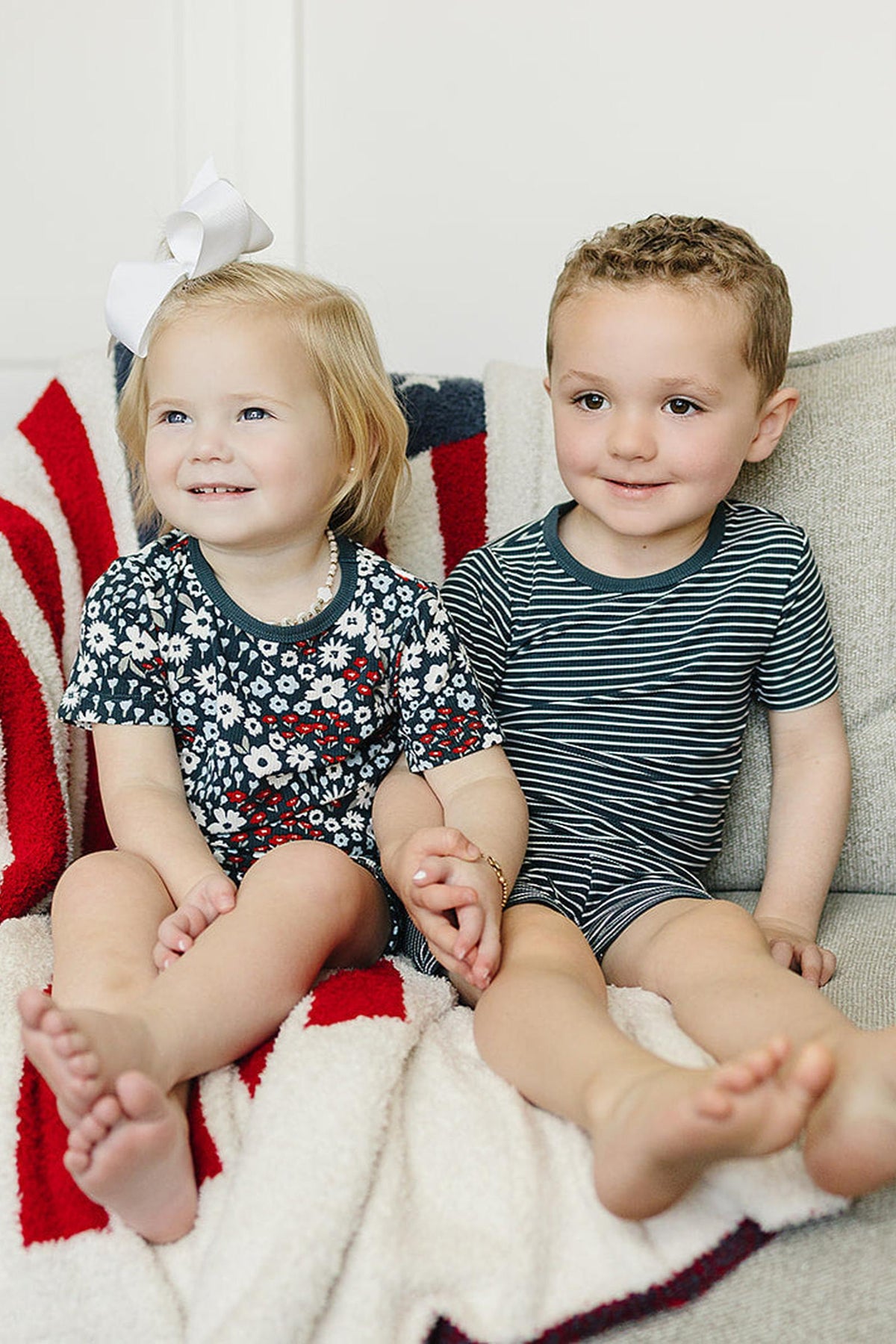 Two toddlers sit on a couch, one wearing a floral outfit, the other in navy striped clothing, holding hands.