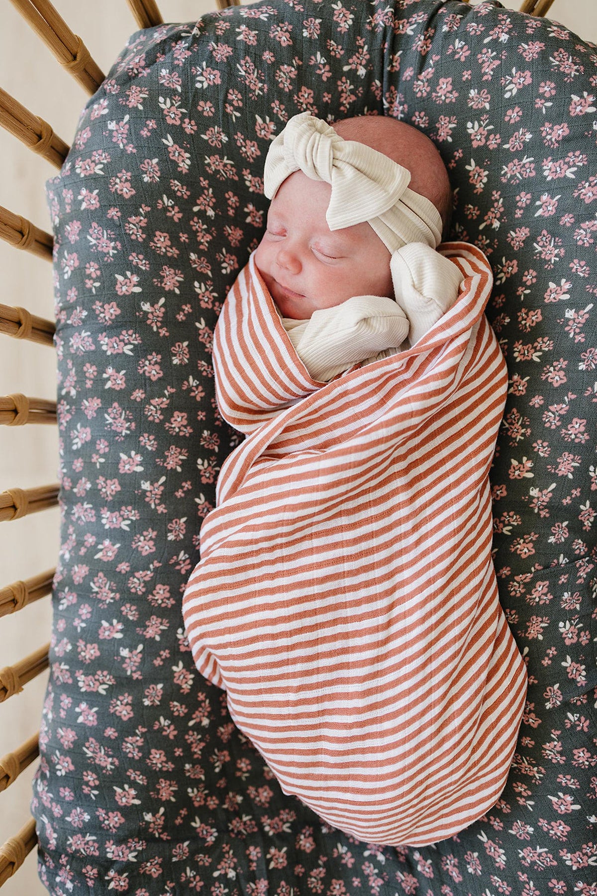 A baby wrapped in a pink striped muslin swaddle blanket, lying on a floral-patterned cushion in a wicker bassinet.