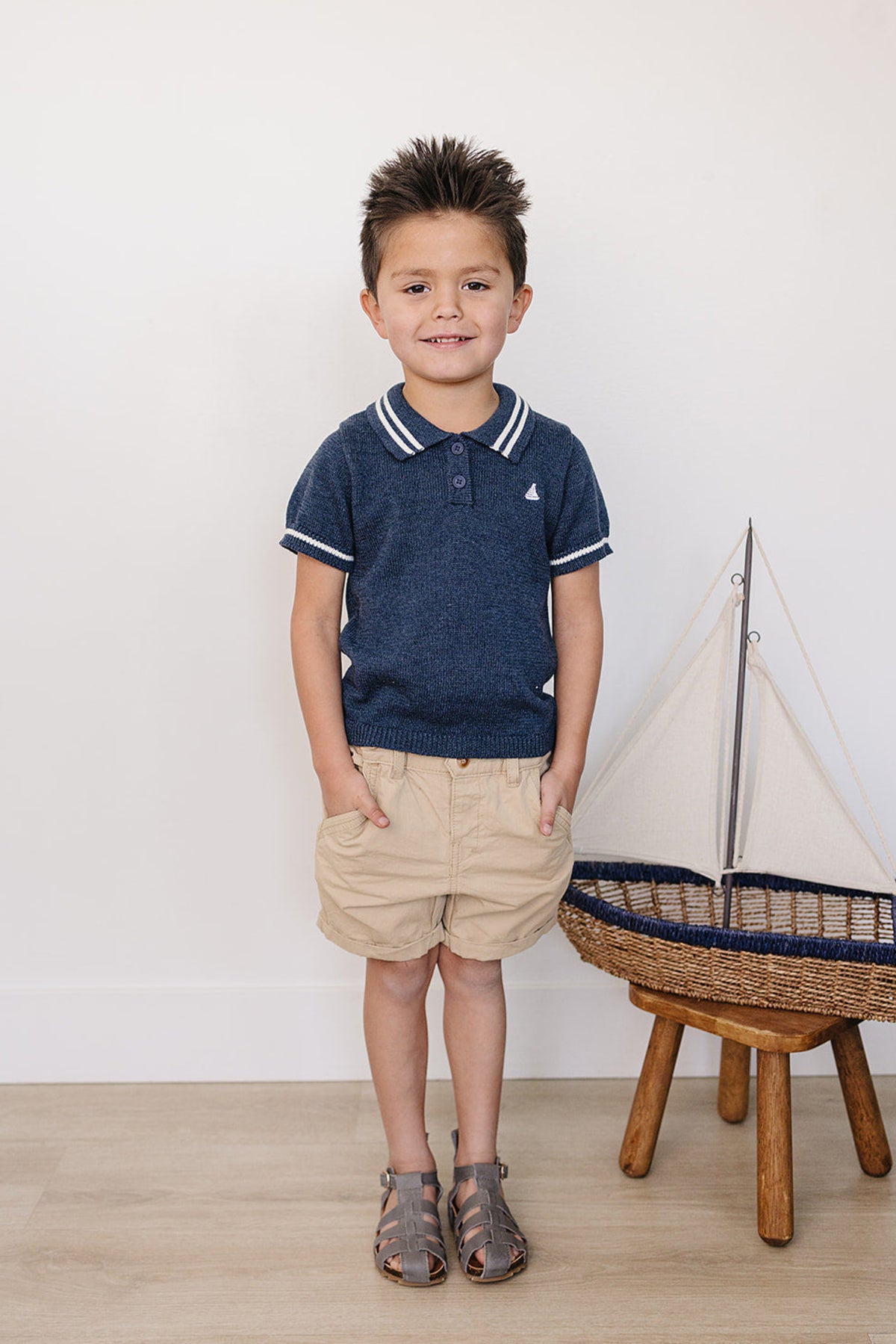 A young boy stands smiling in a navy collared knit shirt and beige shorts, next to a decorative sailboat.