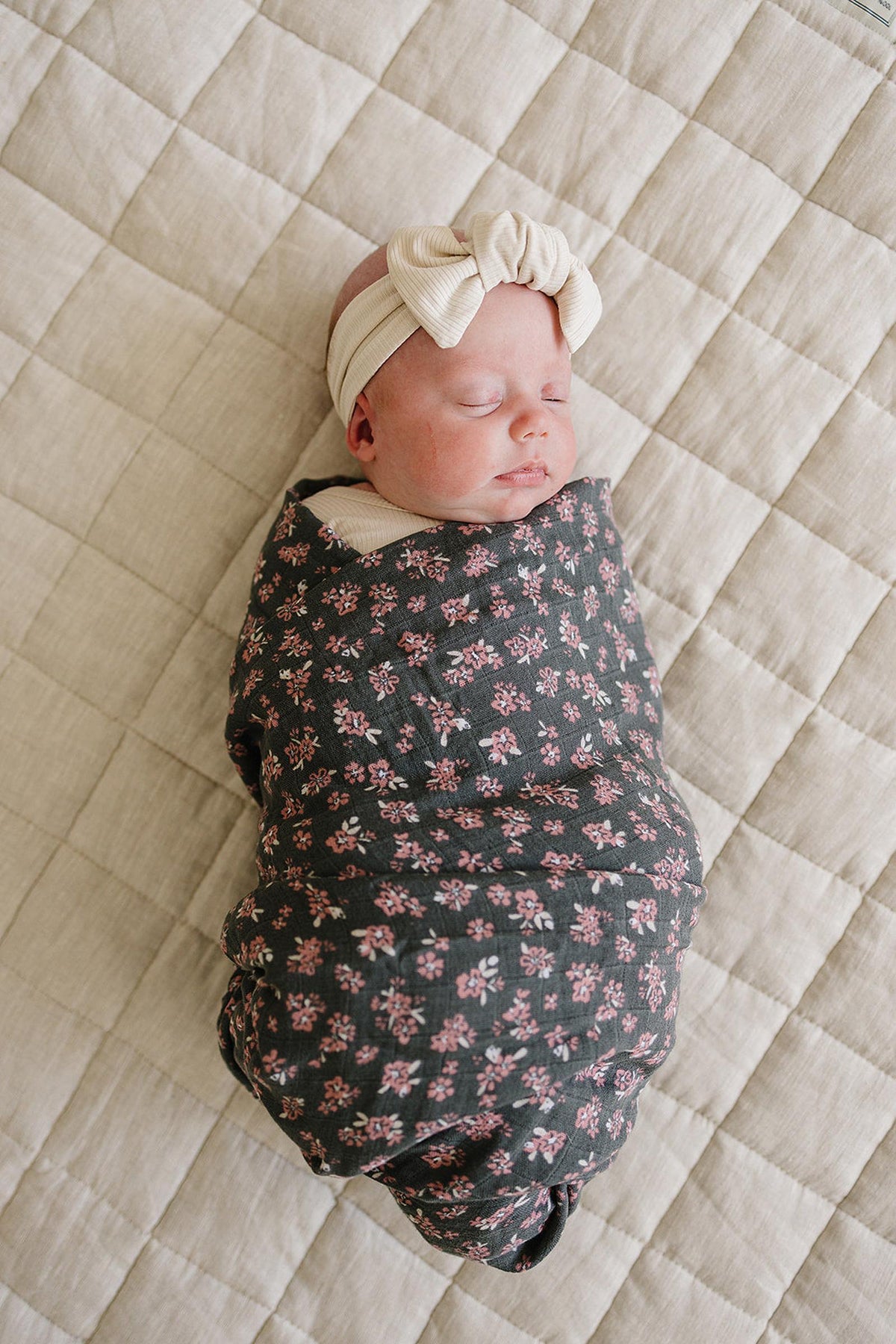 A baby sleeps wrapped in a floral-patterned muslin swaddle blanket, wearing a cream headband on a quilted surface.