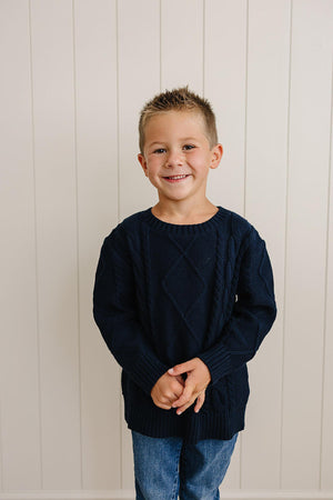A smiling child wearing a navy cable knit sweater and blue jeans stands against a light-colored paneled wall.