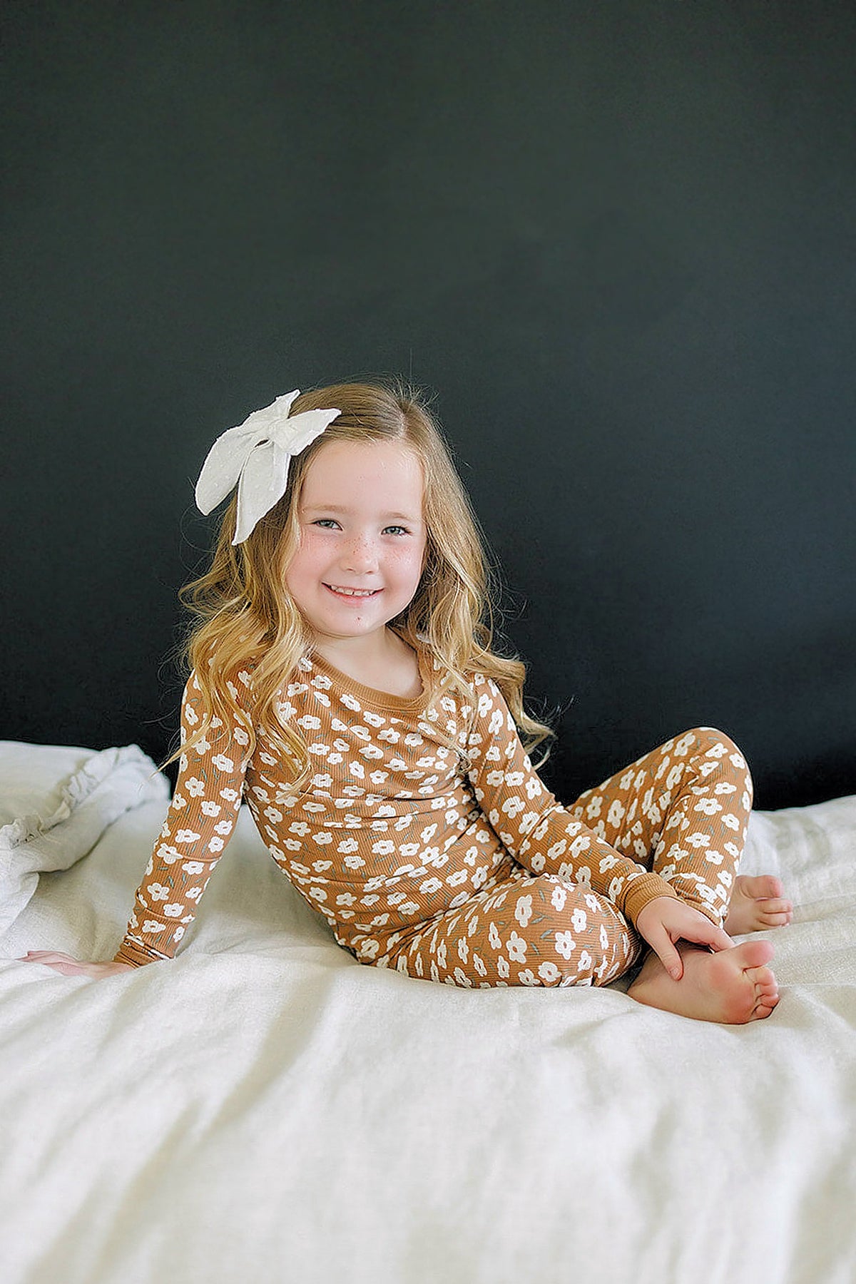 A smiling girl sits on a bed, wearing a mustard floral ribbed pajama set with long sleeves and matching pants.