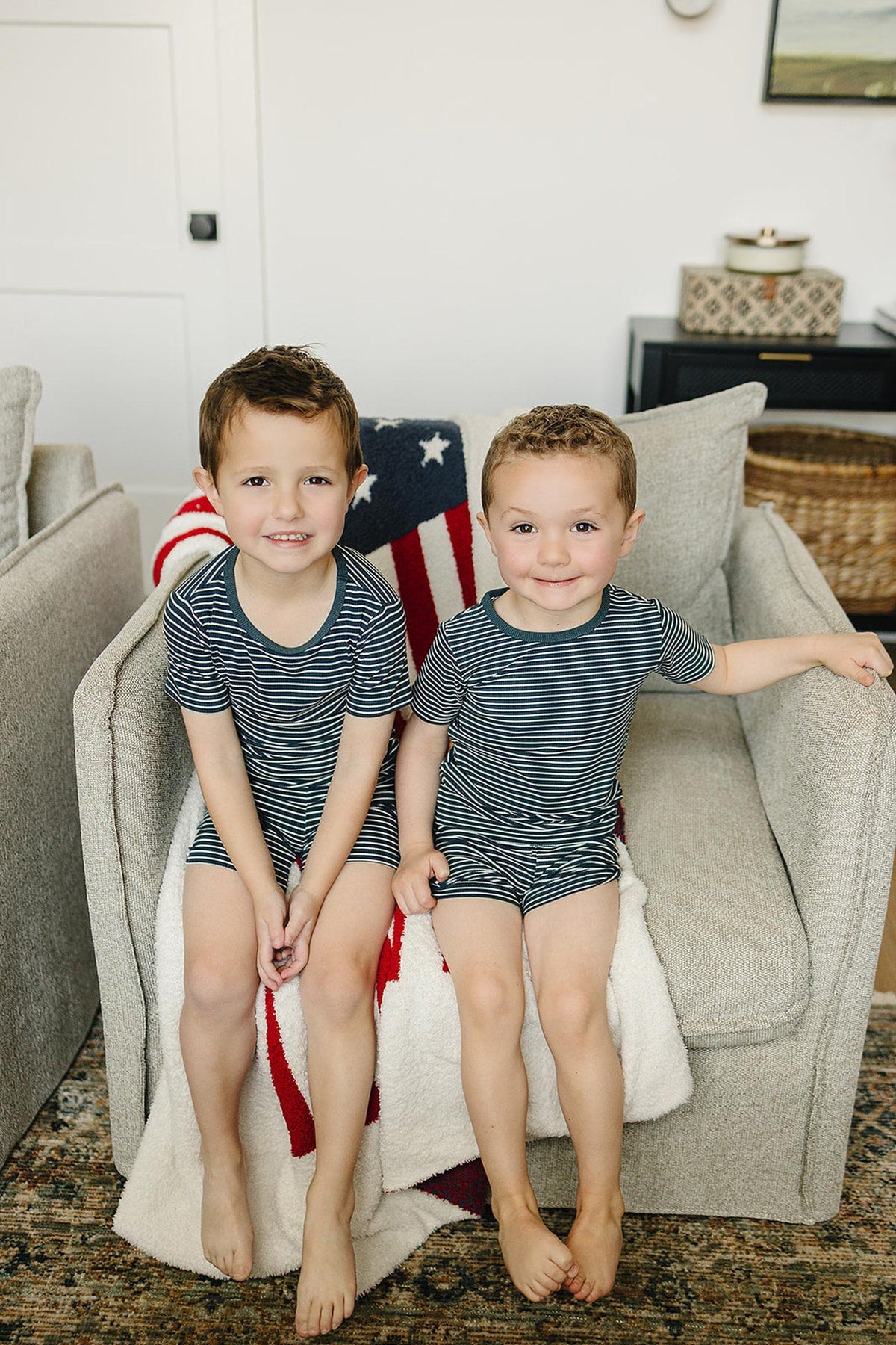 Two children sit on a sofa wearing navy striped shorts sets, with a patriotic blanket draped behind them.