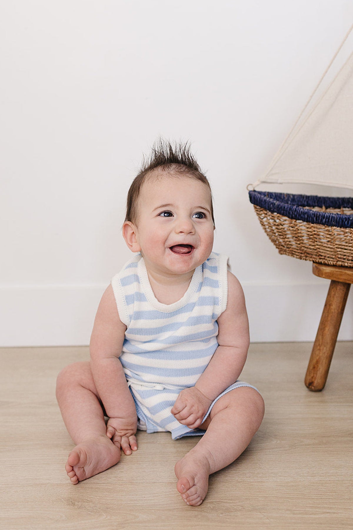 A smiling baby sits on a light wooden floor, wearing a blue and white striped tank outfit.