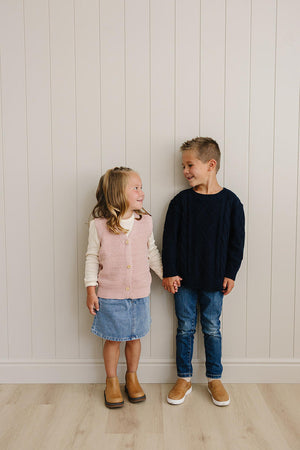 Two children smiling and holding hands; one wears a pink knit vest and the other a navy sweater.
