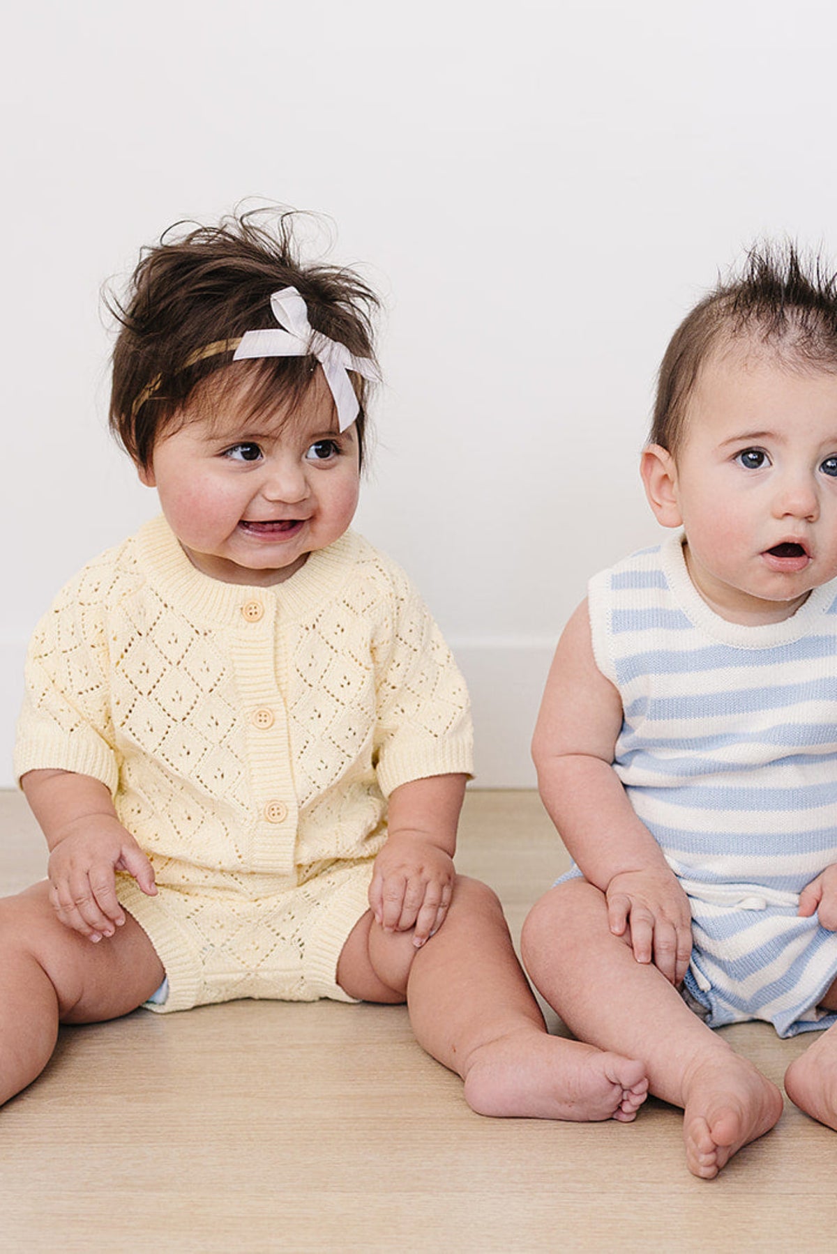 Two babies sit on a light wooden floor; one wears a soft yellow knitted cardigan set with buttons, while the other is in a striped light blue romper.