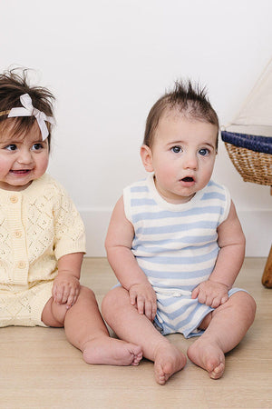 A toddler in a blue and white striped tank set sits next to a baby wearing a yellow knit outfit,.