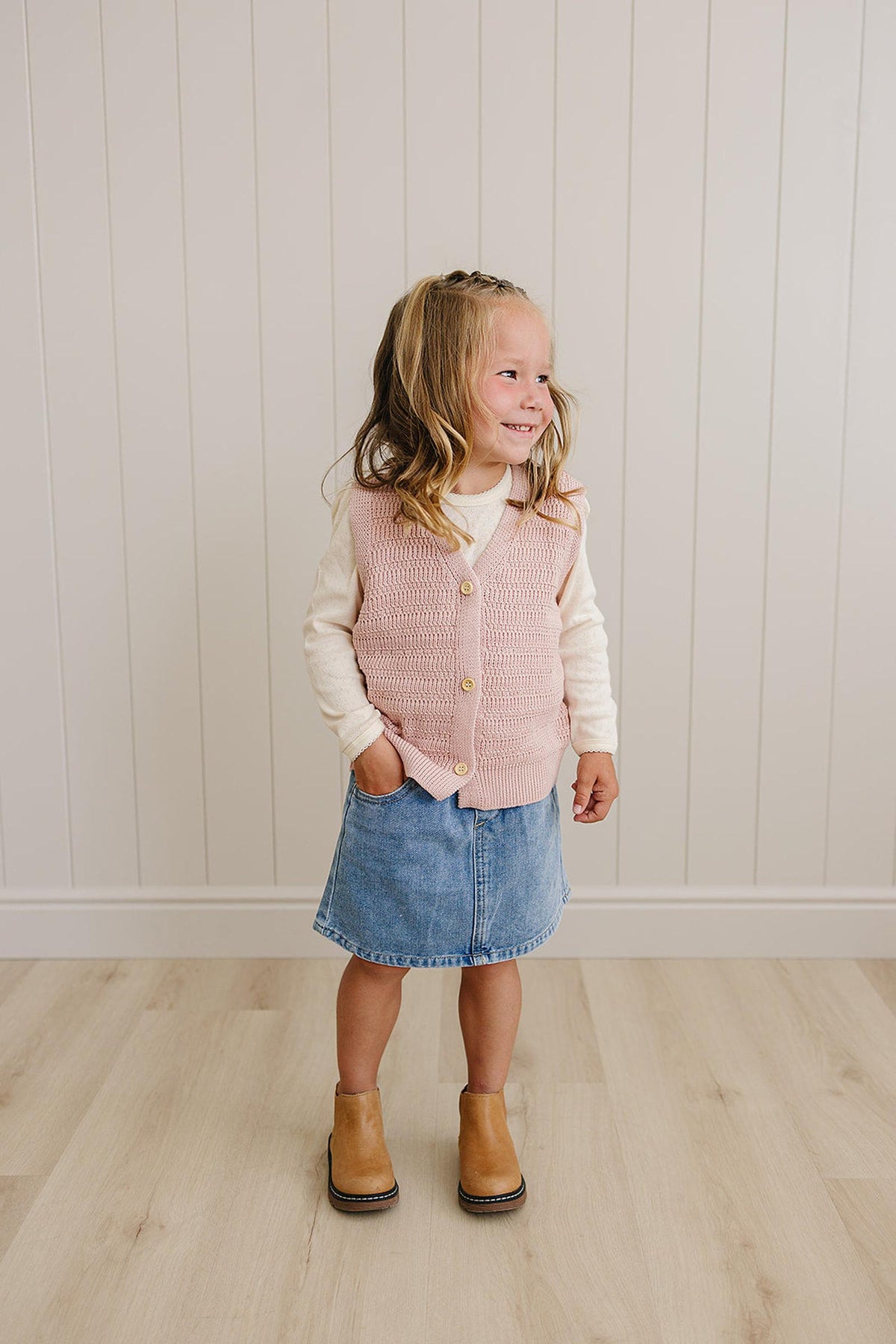 A child wearing a pink knit button-down vest over a cream shirt, denim skirt, and brown boots indoors.