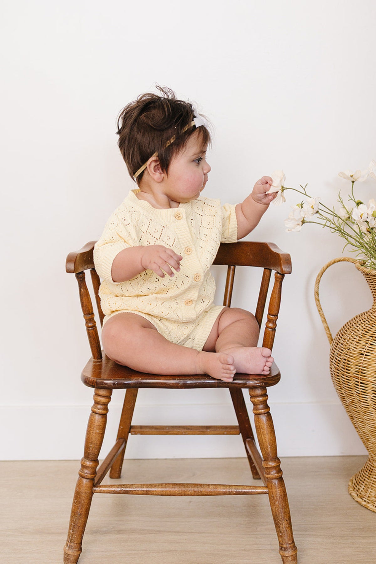 A baby sits on a wooden chair, wearing a soft, yellow knitted cardigan and matching bloomer set. The child is playfully reaching for a flower, surrounded by a neutral backdrop.