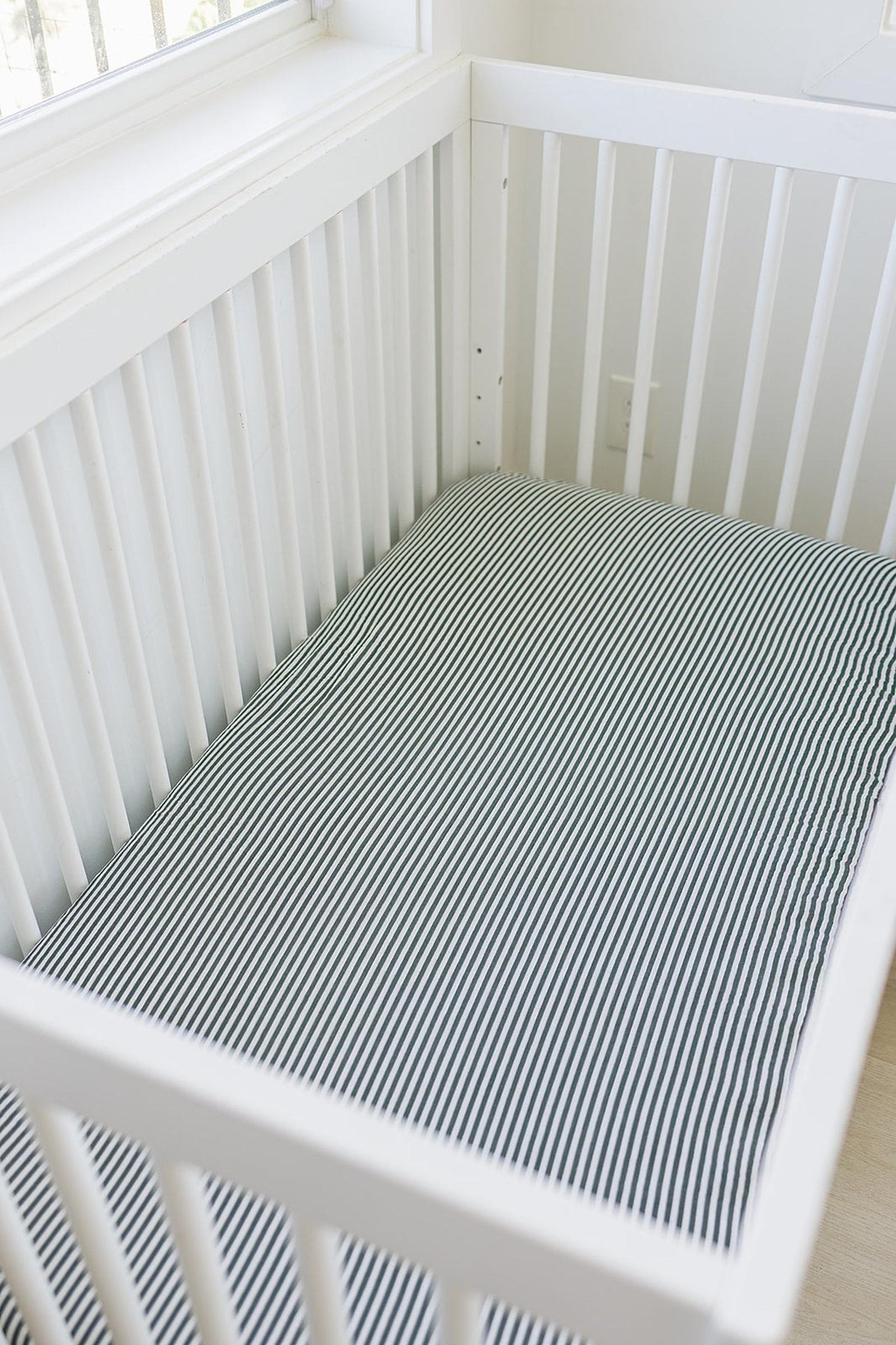 White crib with a green striped muslin sheet, placed in a bright room near a window.