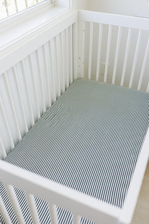 White crib with a green striped muslin sheet, placed in a bright room near a window.
