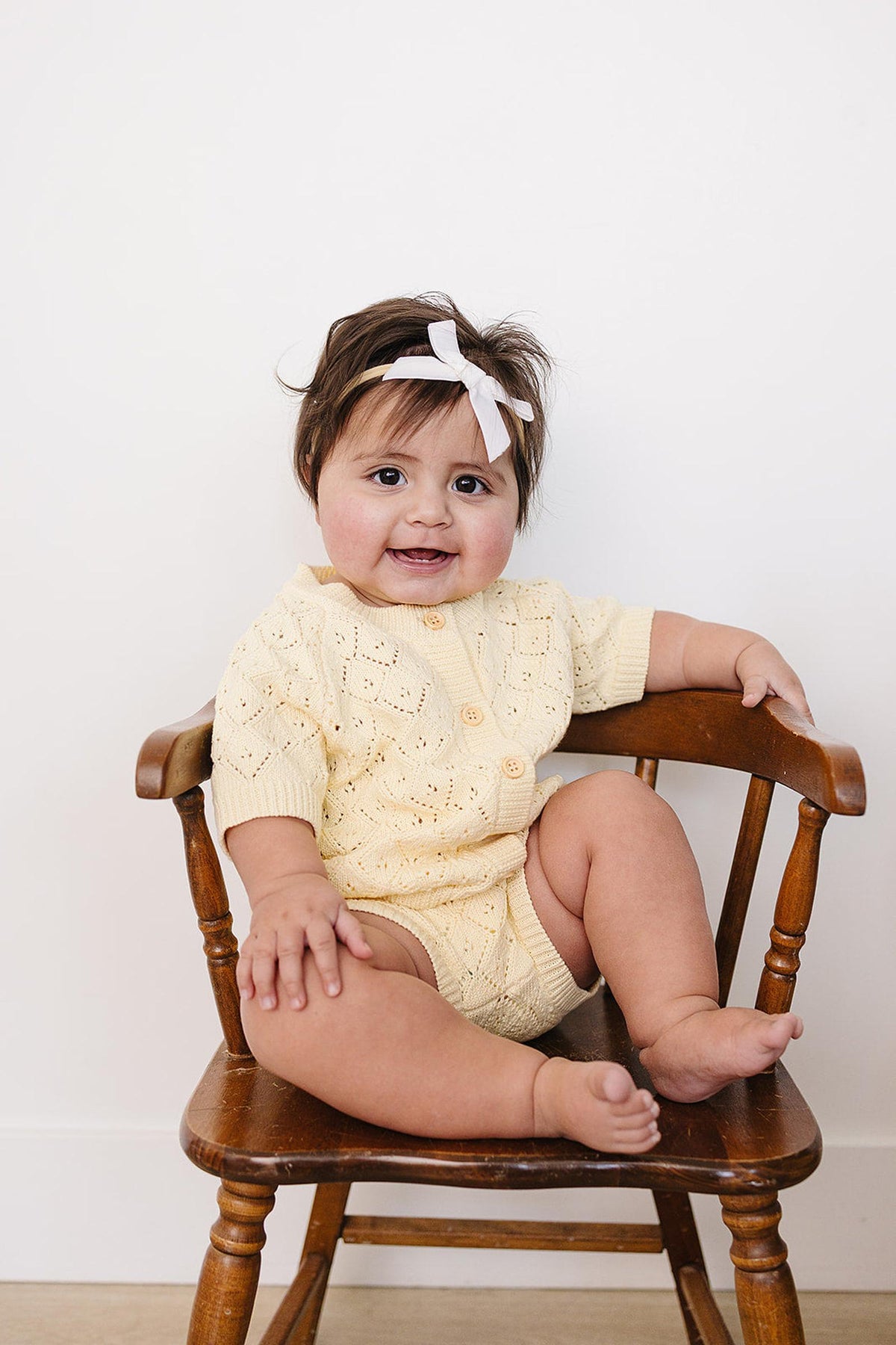A happy baby girl sits on a wooden chair, wearing a soft yellow cardigan and bloomer set. She has a small white bow in her hair, adding a charming touch.