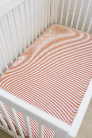 White crib with a pink striped muslin sheet, offering a cozy and stylish look for a baby nursery.