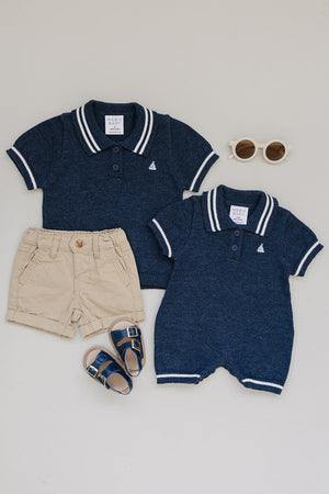 Two navy collared knit shirts, beige shorts, navy sandals, and white sunglasses arranged on a light background.