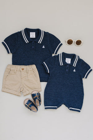 Two navy collared knit rompers and a pair of beige shorts are arranged with navy sandals and round sunglasses.