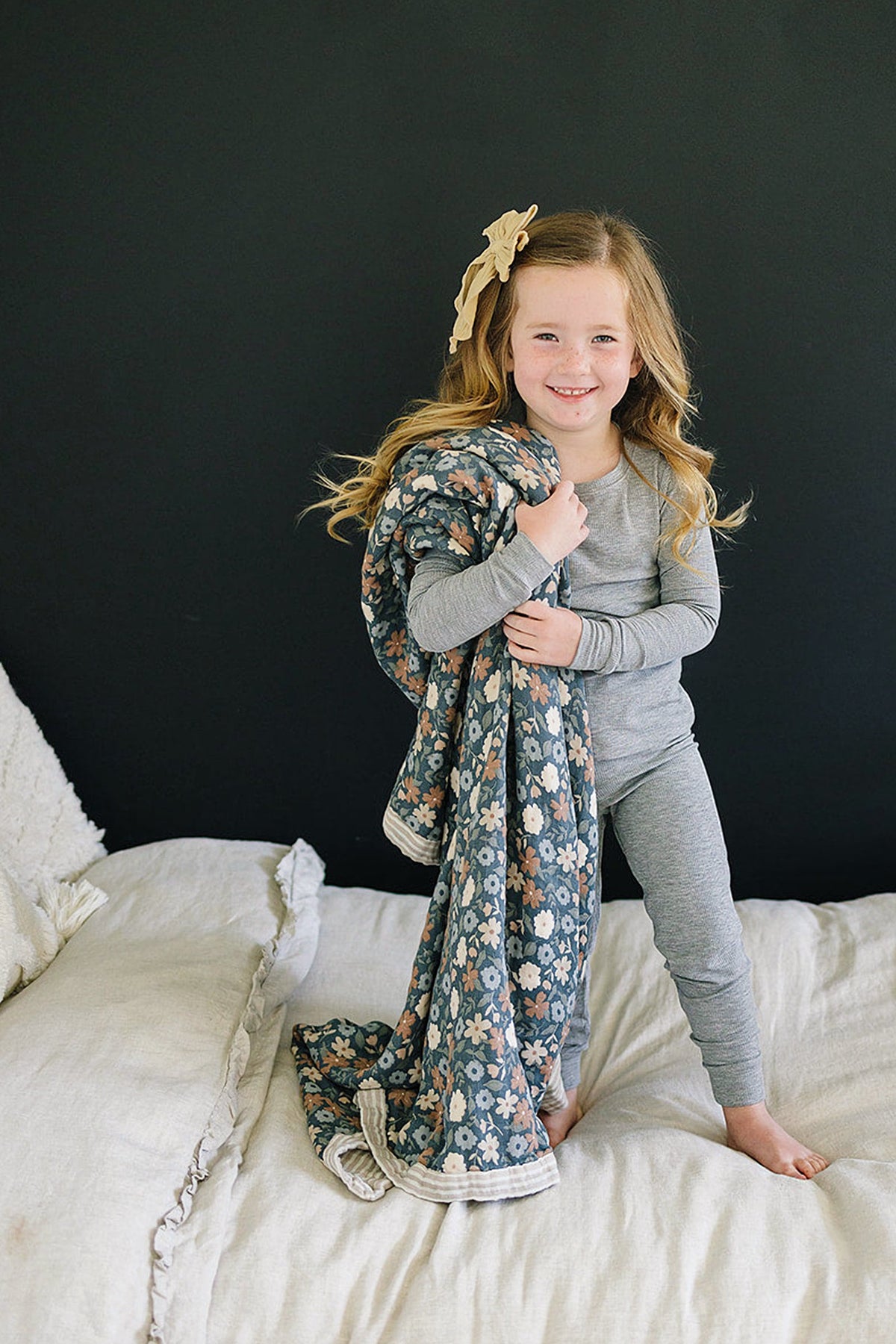 A child in a grey pajama set holds a floral patterned blanket, standing on a bed with white bedding.