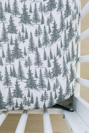White crib sheet with green forest tree pattern, fitted on a crib with white wooden slats.
