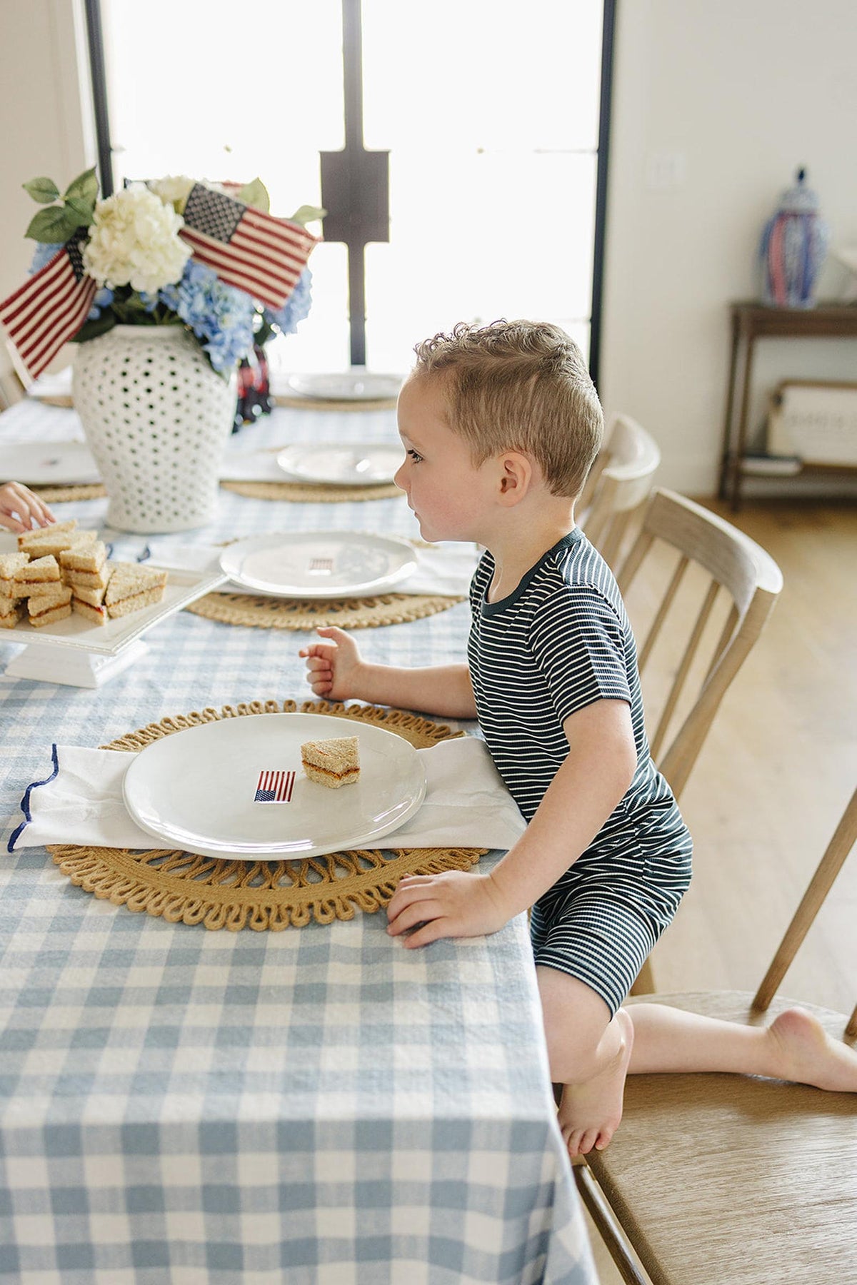 A child in a navy striped outfit sits at a table with American flag-themed decor and sandwiches.
