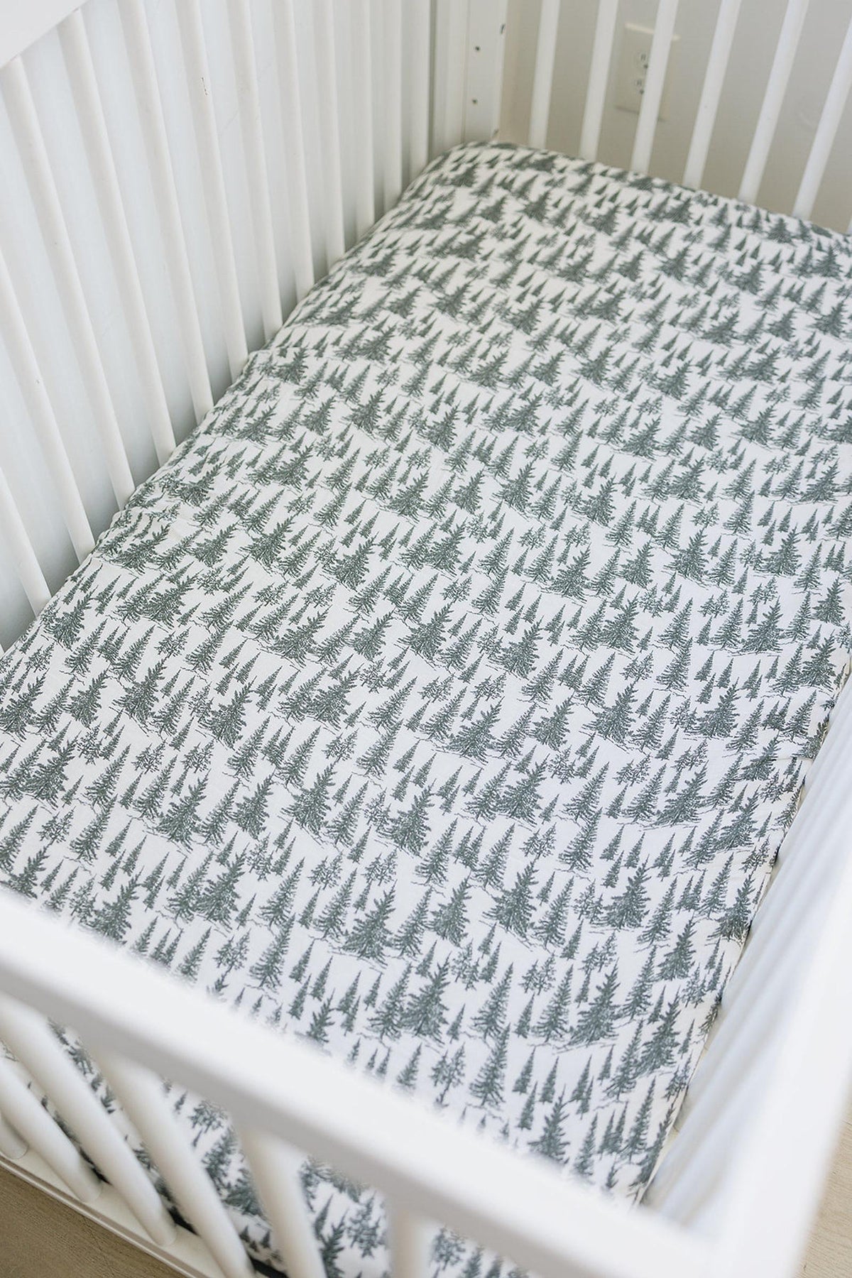 White crib with a forest-patterned muslin sheet featuring green trees, creating a cozy and natural nursery setting.