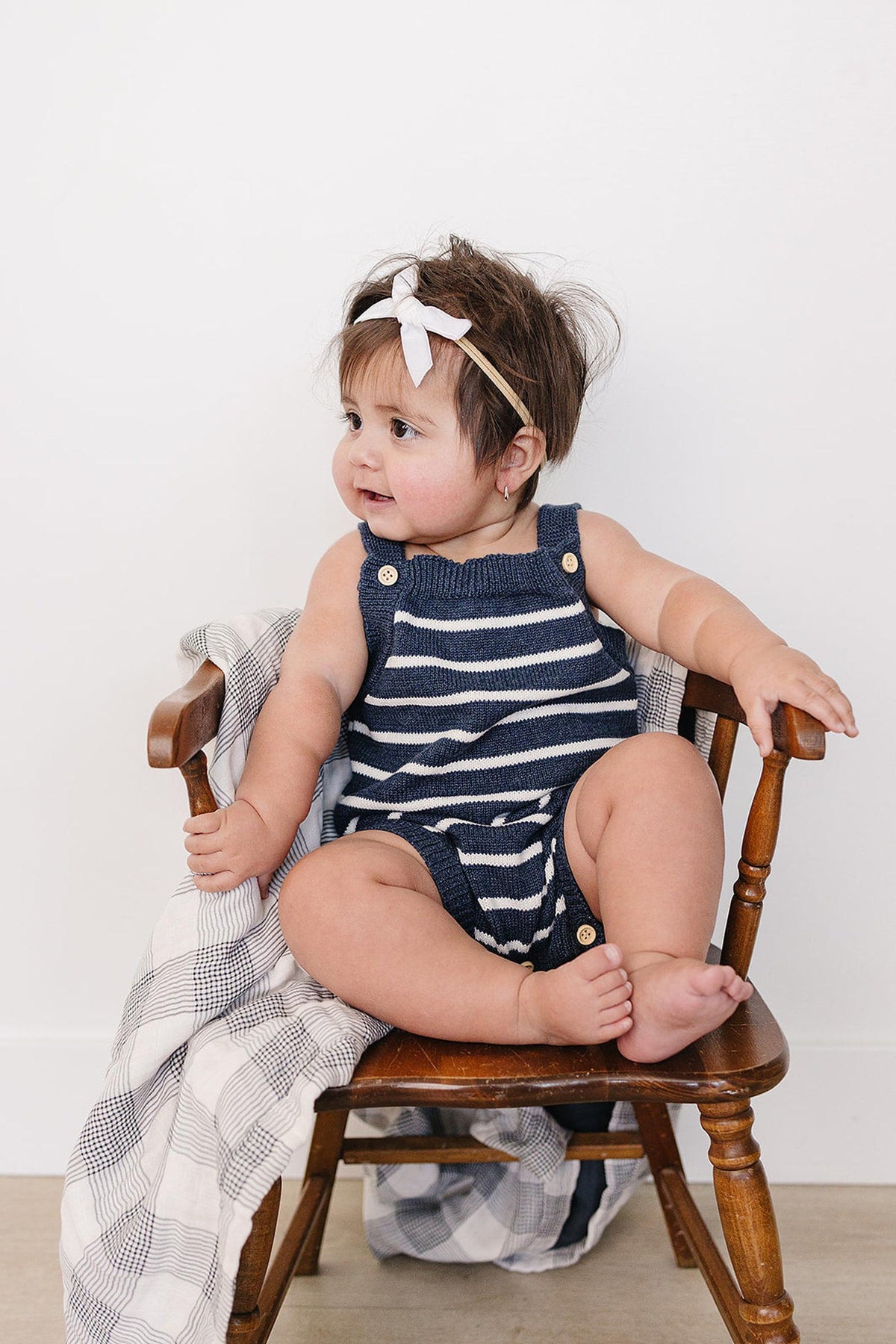 A baby sits on a wooden chair wearing a navy and white striped knit bubble romper, featuring snap closures and a stylish headband, with a cozy blanket draped beside them.