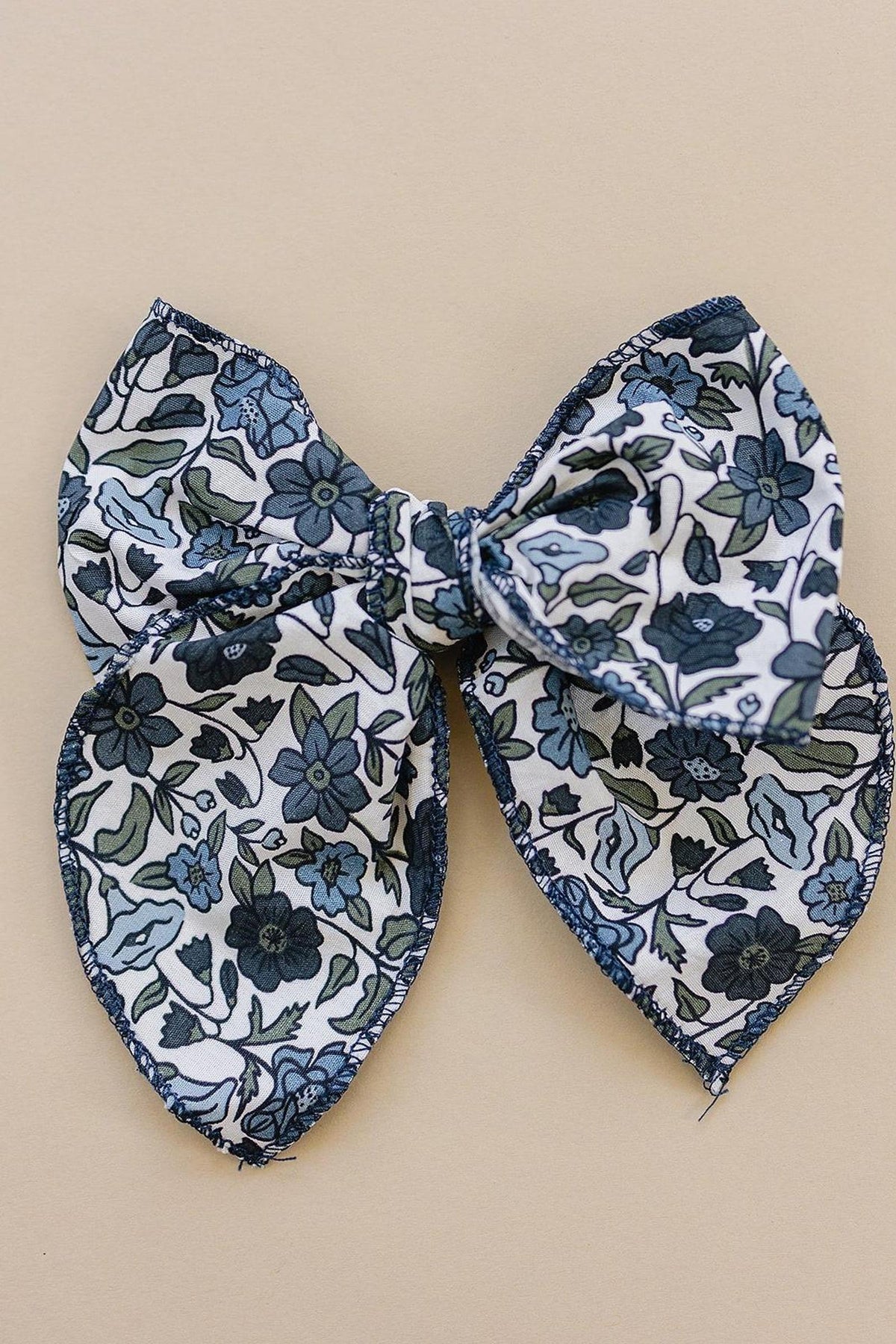 Floral-patterned fabric bow with blue and green hues on a beige background, handcrafted for baby and toddler wear.