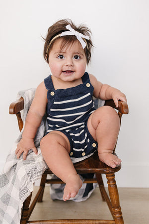 A cute baby girl sits on a wooden chair, wearing a navy and white striped knit bubble romper with snap closures.