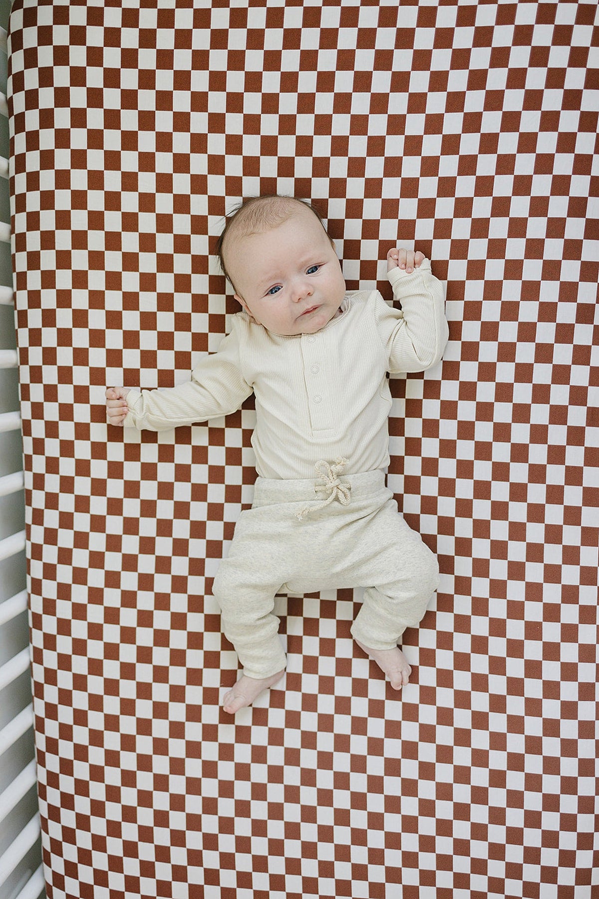 A baby lies on a rust checkered crib sheet, wearing a cream outfit, in a white crib.
