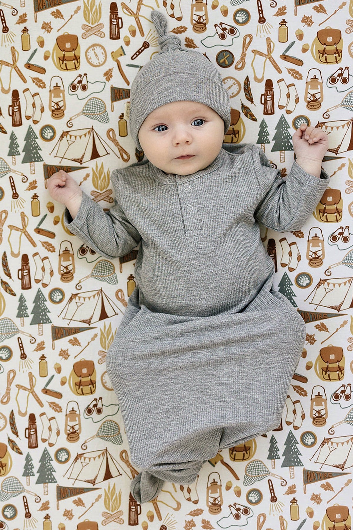 A baby in a grey ribbed knot gown and matching hat lies on a nature-themed patterned blanket.