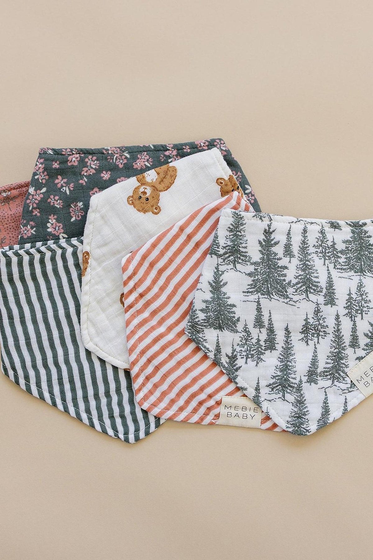 Five Mebie Baby muslin bibs with various patterns, including stripes, bears, and trees, displayed on a beige surface.