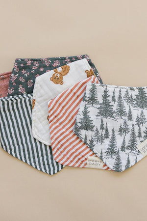 Five Mebie Baby muslin bibs with various patterns, including stripes, bears, and trees, displayed on a beige surface.