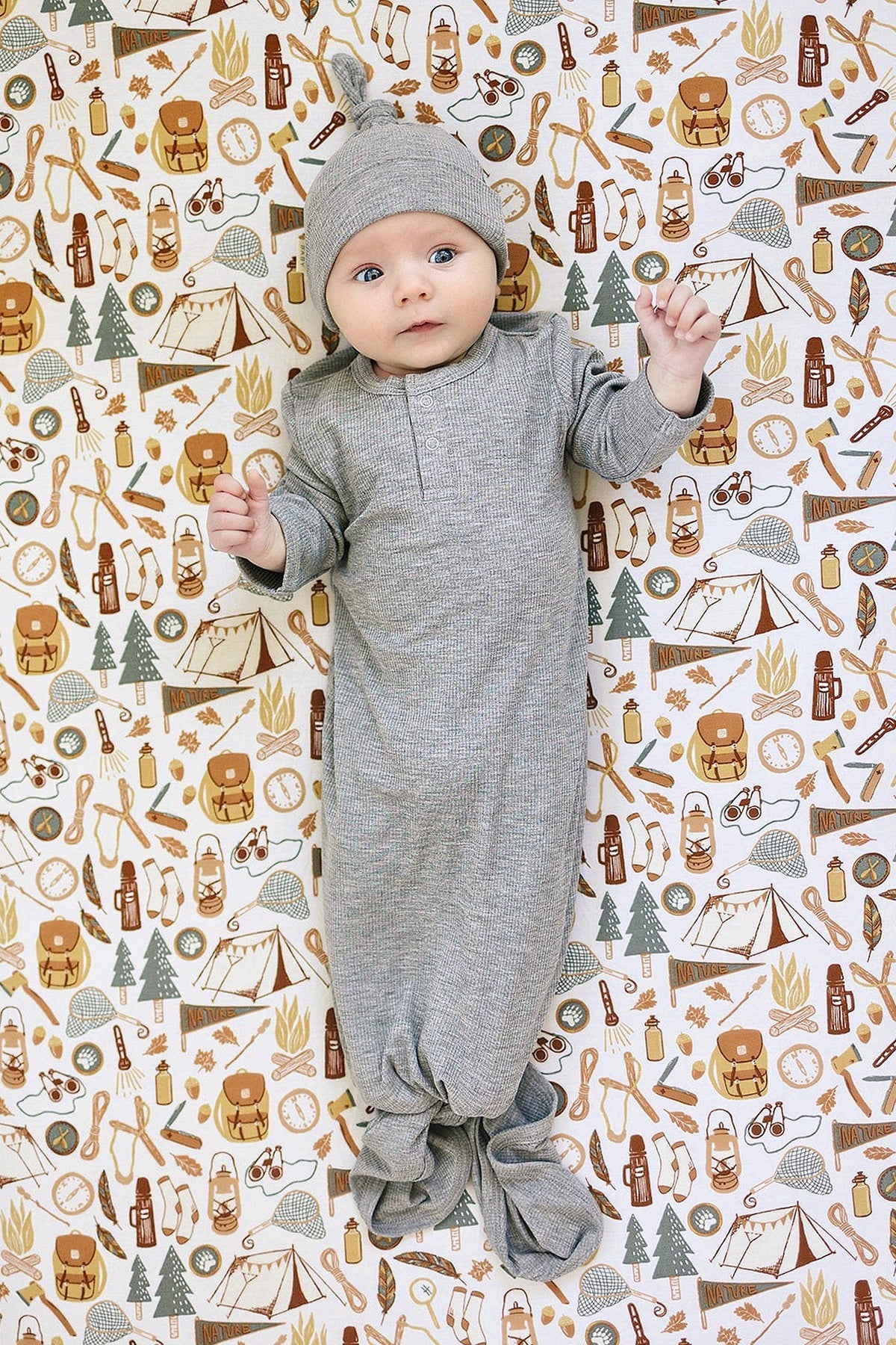 Baby in a heather grey ribbed gown and matching hat on a camping-themed patterned background with nature illustrations.