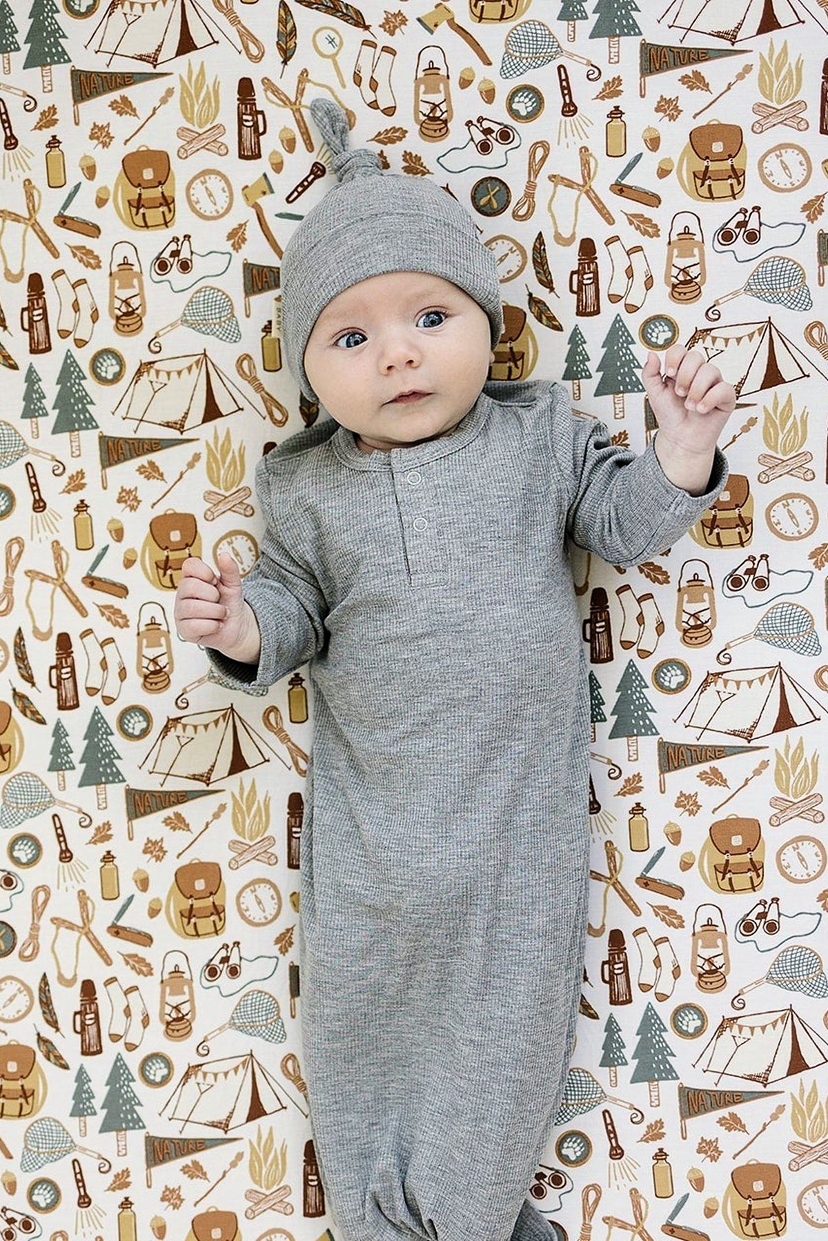 Baby in a gray outfit lies on a camping-themed crib sheet with tents, trees, and outdoor gear illustrations.