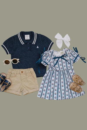 A navy polo shirt, beige shorts, and a blue floral block print dress are arranged with matching sandals and sunglasses. A white bow completes the playful outfit for young children.