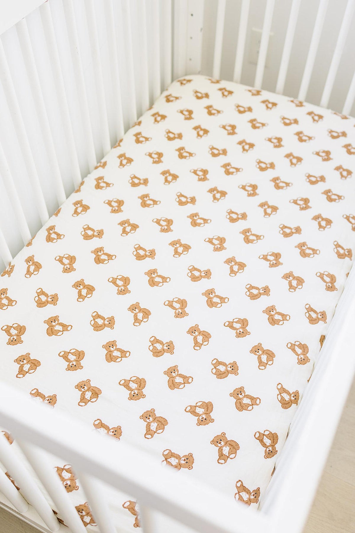 White crib sheet with a brown teddy bear print in a white crib setting. Made from soft muslin cotton.