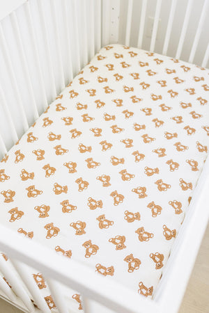 White crib sheet with a brown teddy bear print in a white crib setting. Made from soft muslin cotton.