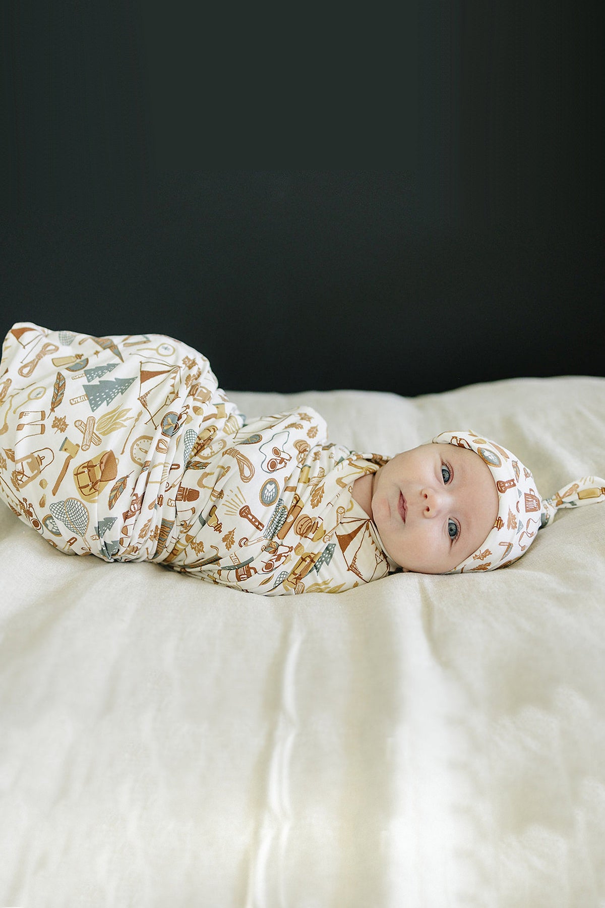 A baby is swaddled in a camping-themed, bamboo fabric blanket with matching hat, lying on a light bed.