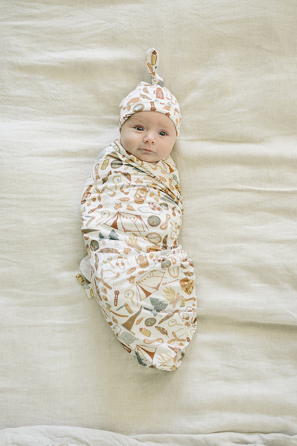 A baby wrapped in a camping-themed swaddle blanket with matching hat on a beige background.