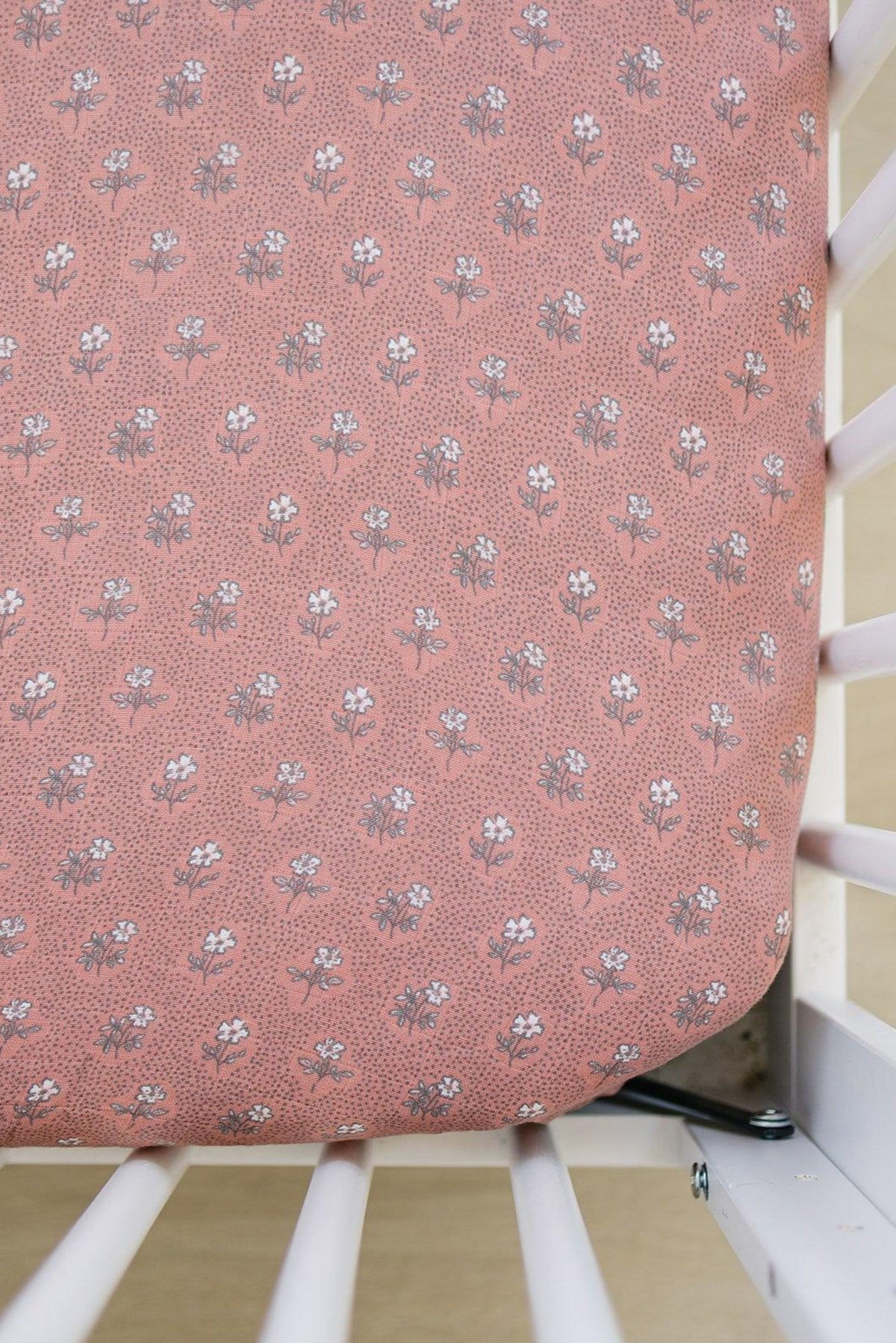 Pink floral muslin crib sheet with a delicate white flower pattern on a crib. Made from soft, breathable fabric.