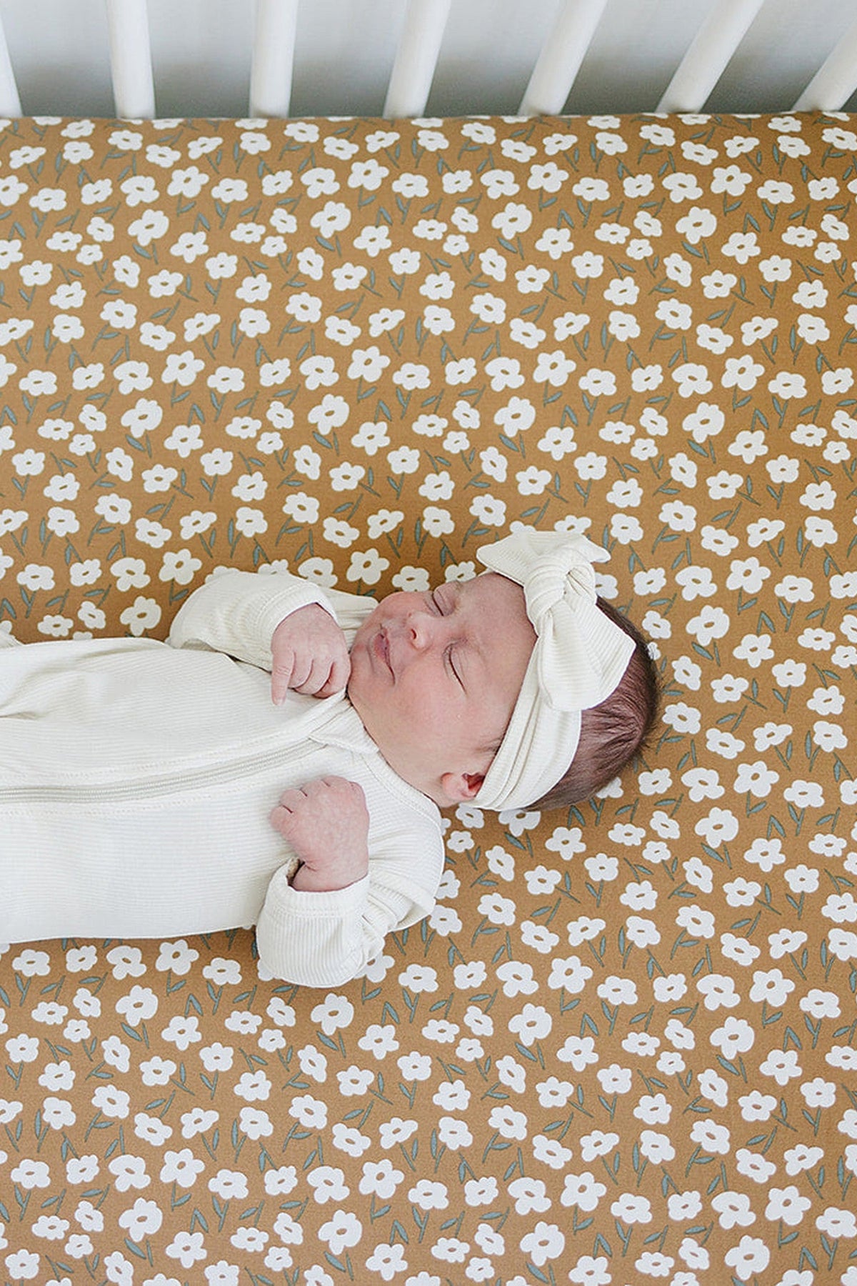 A baby wearing a white outfit and headband lies on a mustard floral crib sheet with white flowers.