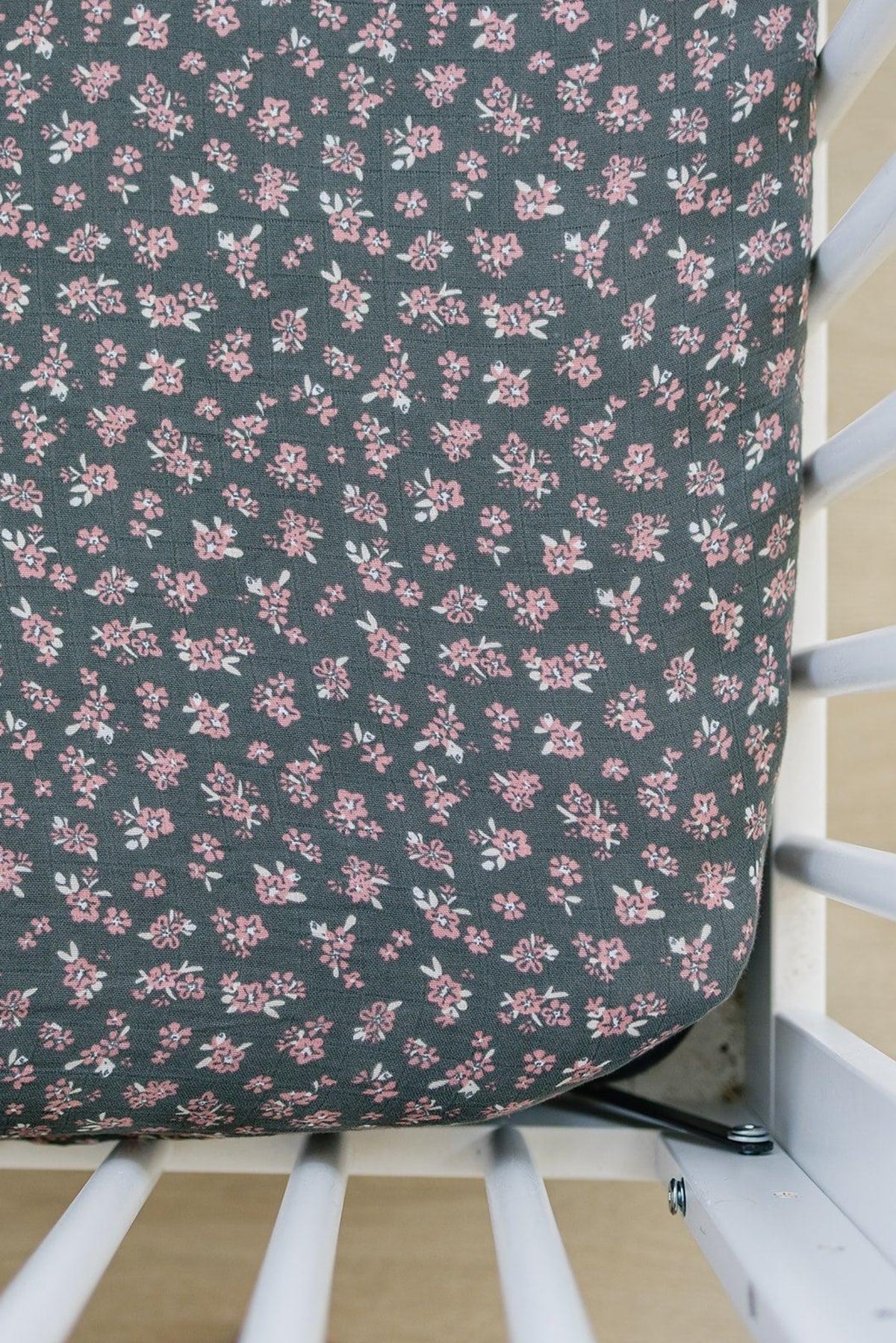 A crib with a dark muslin sheet featuring a pink floral pattern, surrounded by white crib bars.