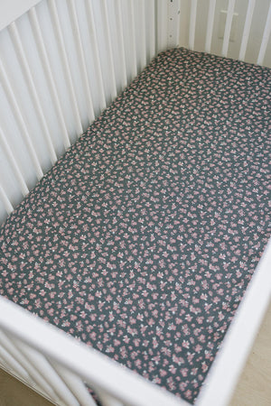 White crib with a dark muslin sheet featuring small pink floral patterns, creating a cozy nursery setting.
