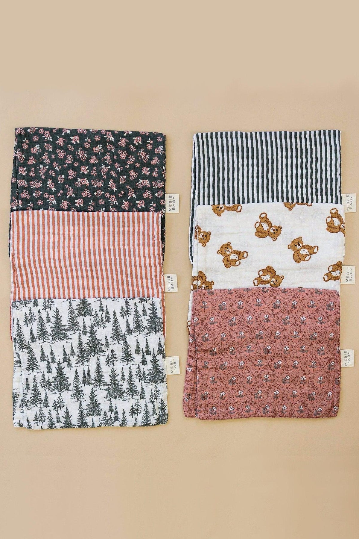 Two sets of Mebie Baby muslin burp cloths with floral, striped, teddy bear, and forest patterns on a beige background.